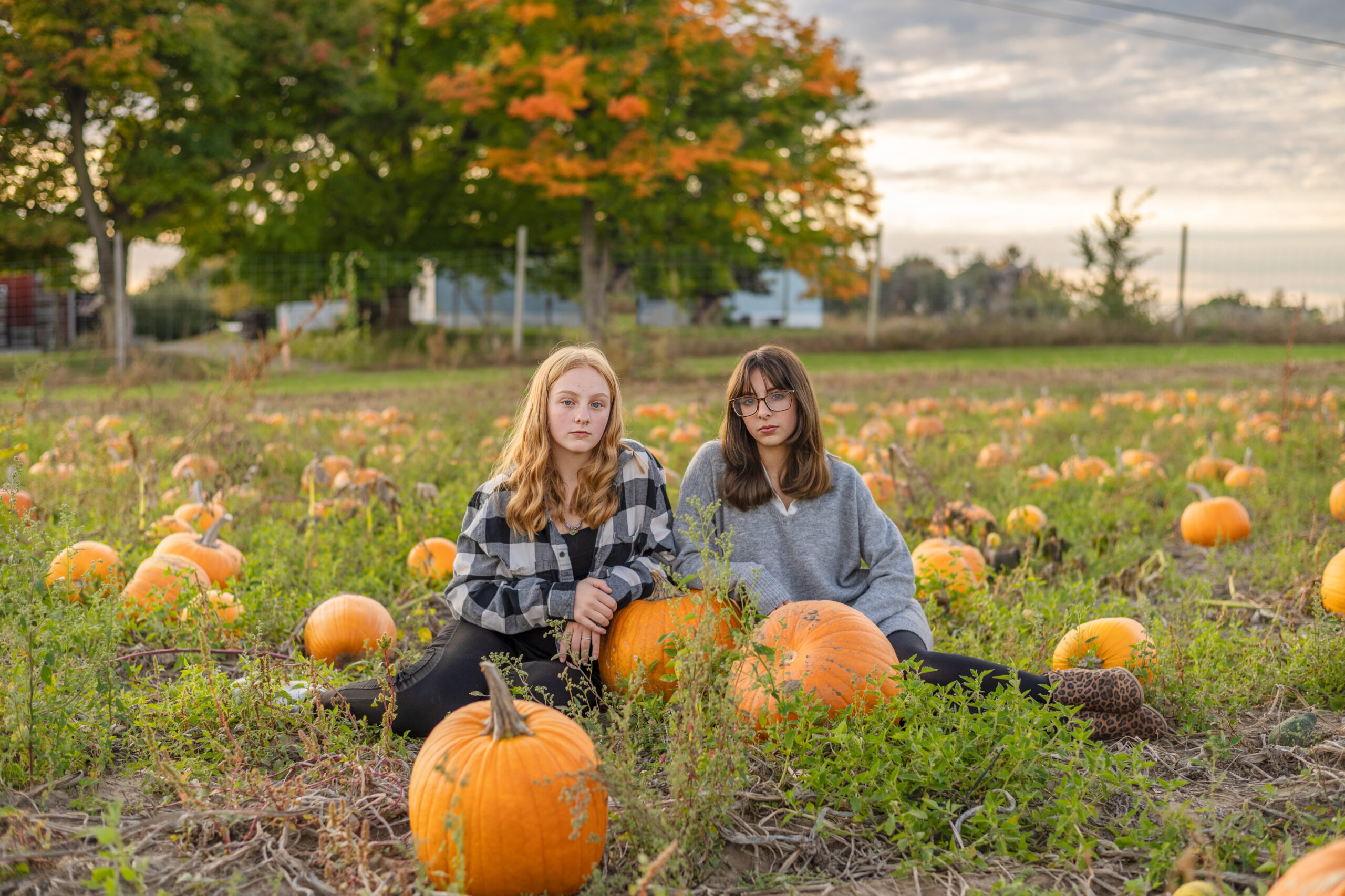 Two teen girls sitting next to each other resting elbows on a pumpkin in local pumpkin patch