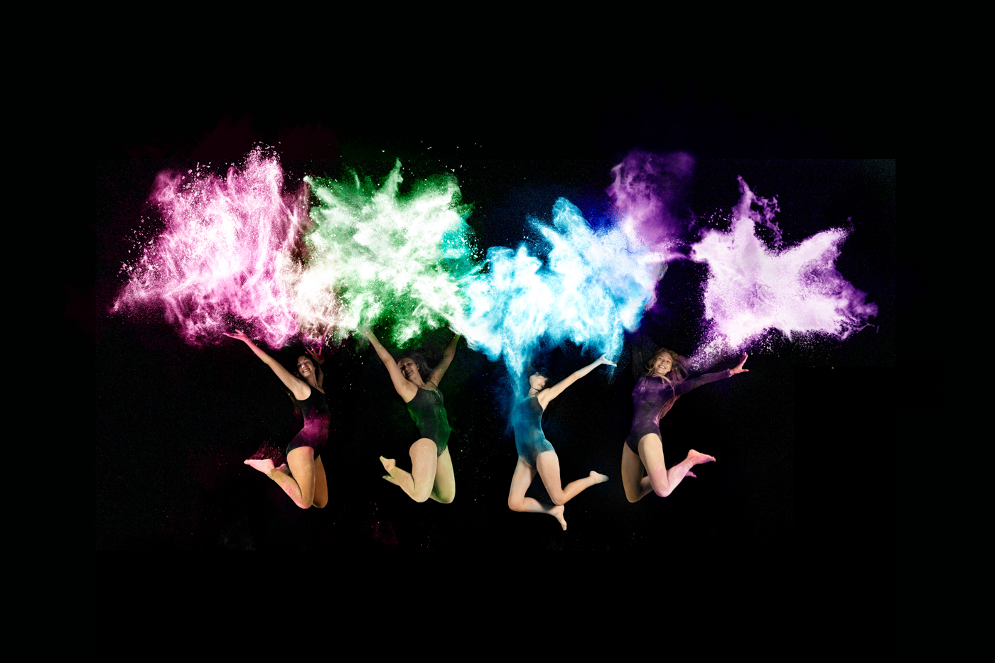 Four dancers in midair, each with colored powder (pink, green, blue, and purple) exploding above them against a black background.