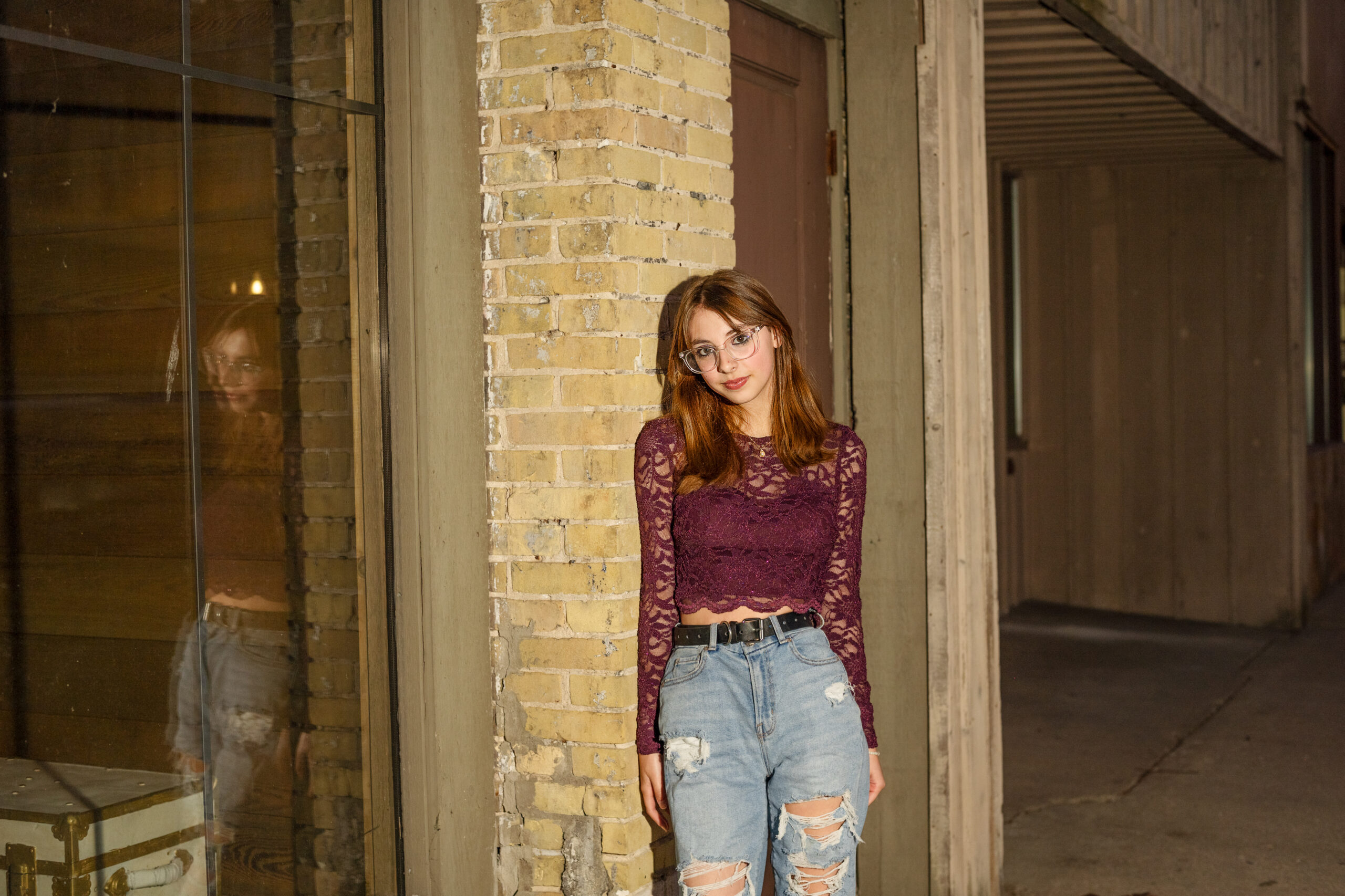 A teenage girl with long hair stands outside next to a brick wall. They are wearing a maroon long-sleeve lace top and ripped jeans.