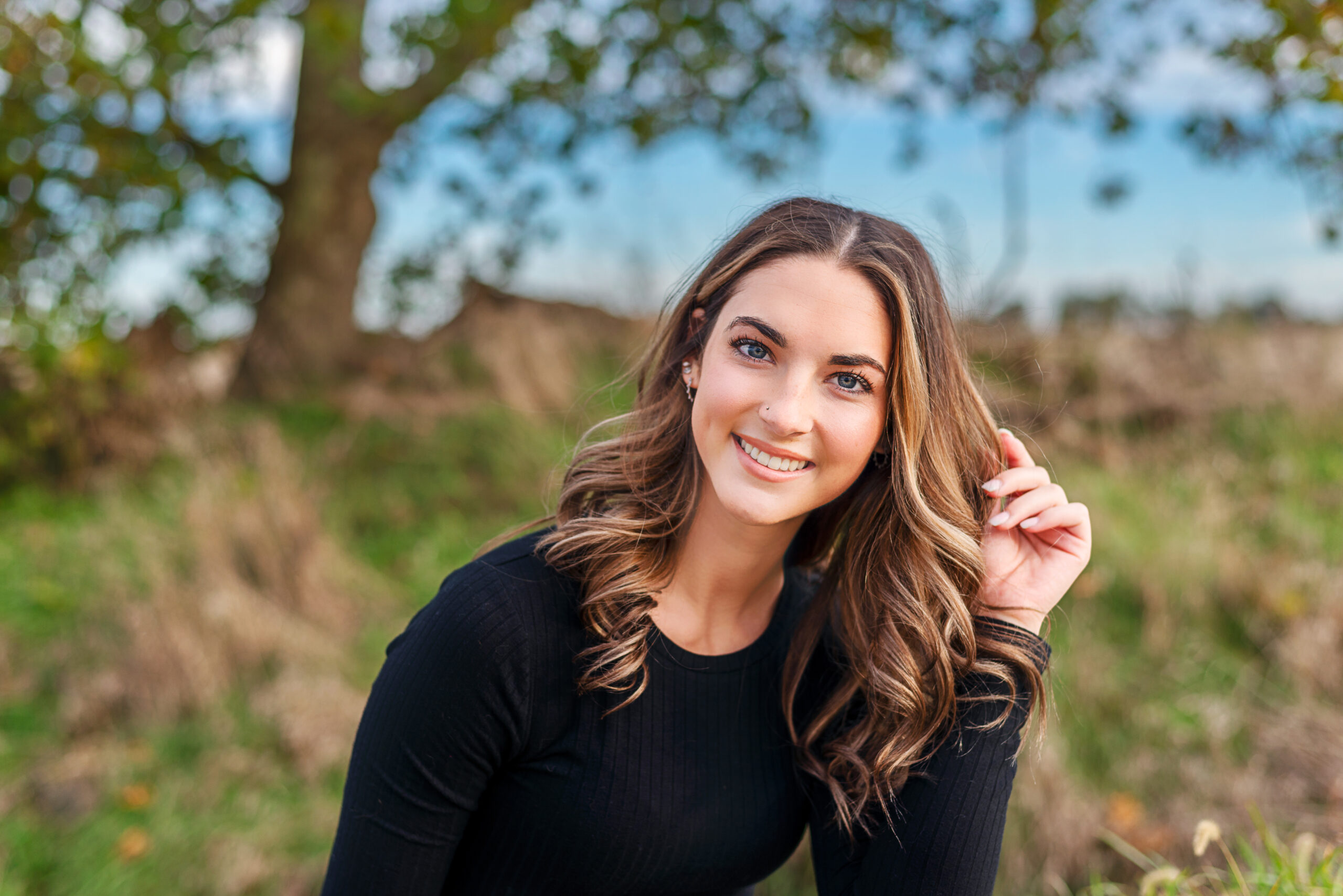 A woman with long wavy hair and a black top smiles at the camera while posing outdoors, with a tree and grassy background.