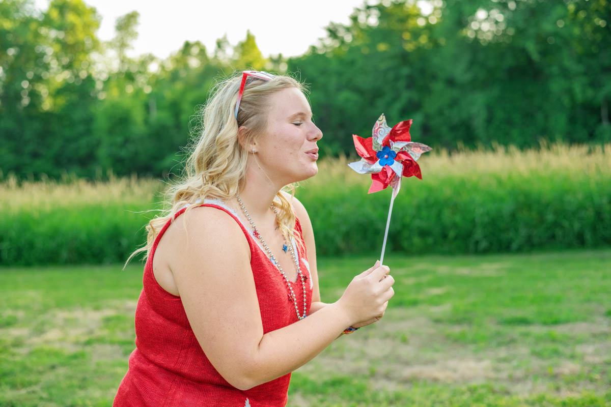 A high school girl in a red sleeveless top stands outdoors, blowing on a red and blue pinwheel. Green trees and grass are in the background.