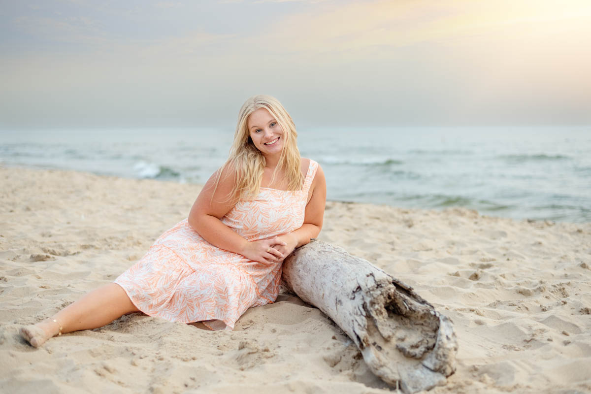 Summer Senior Session A woman in a light dress sits on the sand next to a driftwood log on a beach, with the ocean and a soft sunset sky in the background.