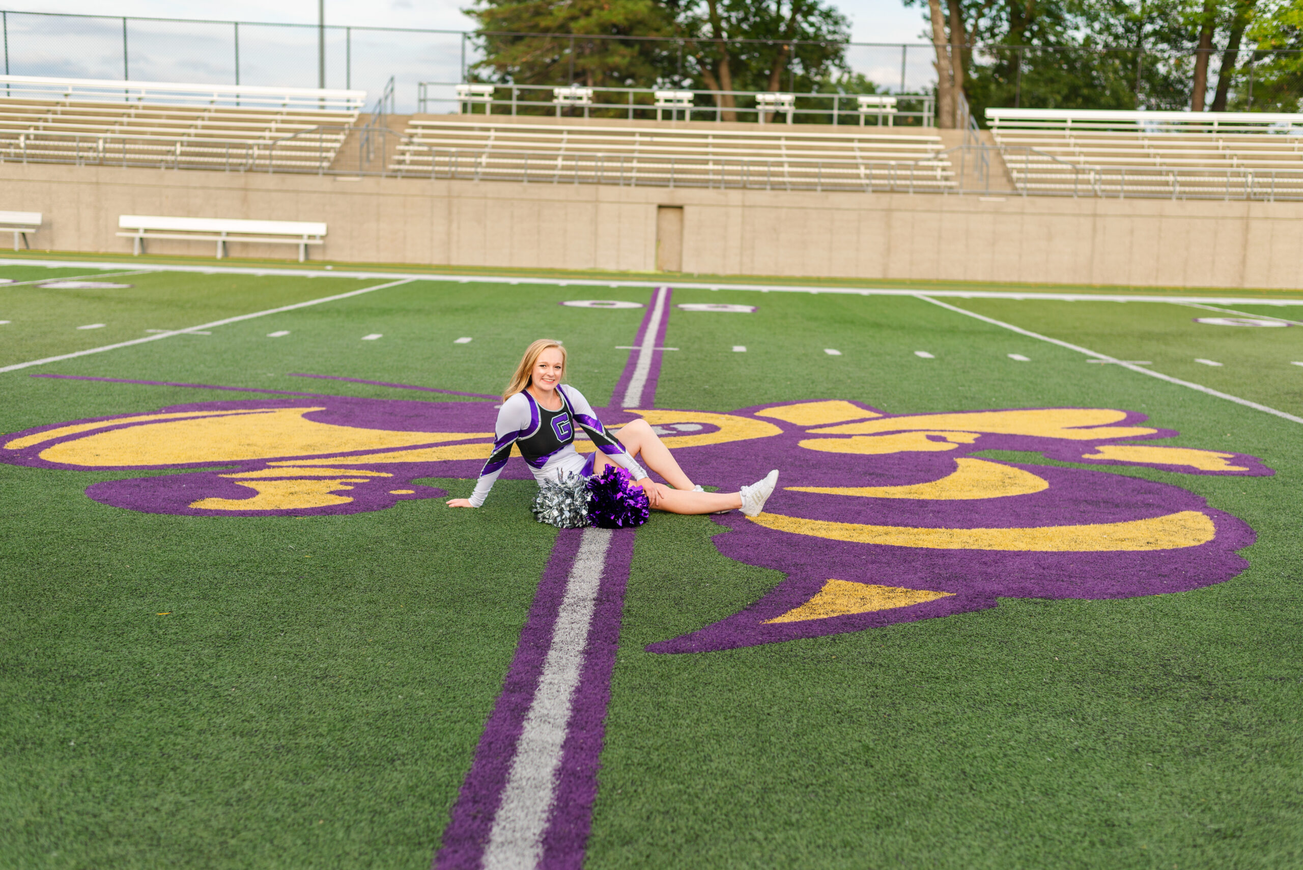 A cheerleader in uniform sits on a large purple and gold logo at the center of a football field.