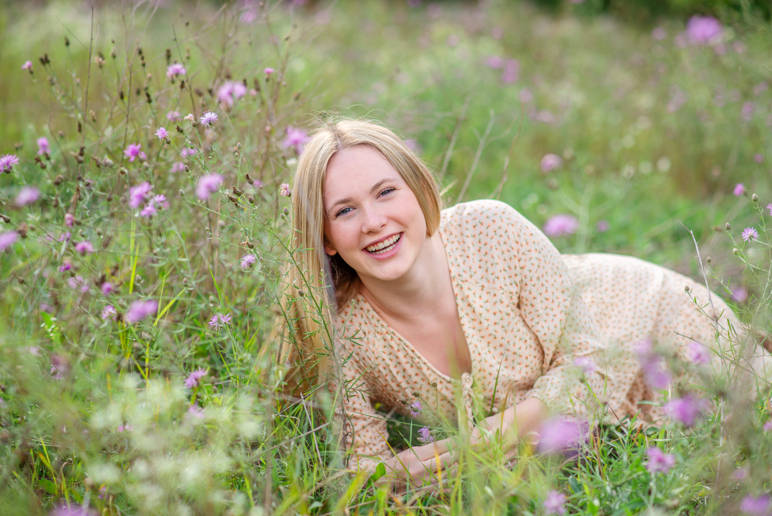 A high school senior girl with long blonde hair, wearing a light-colored dress, smiles while lying in a field of purple wildflowers.