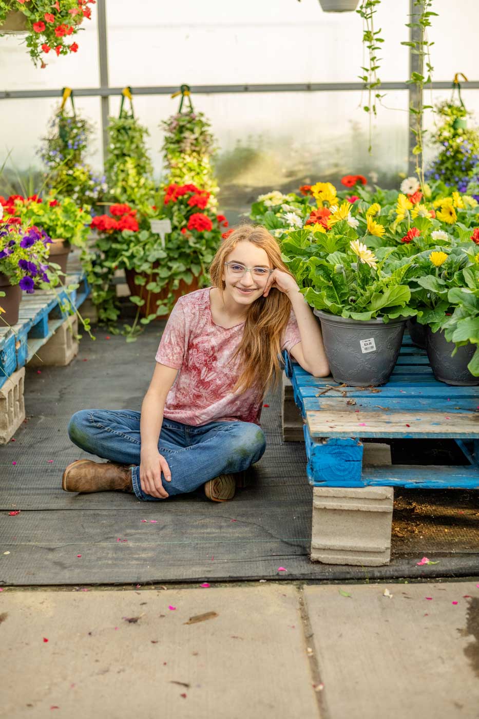 A high school girl with long hair sits on the floor of a greenhouse beside pots of colorful flowers, surrounded by vibrant plants and wooden pallets.