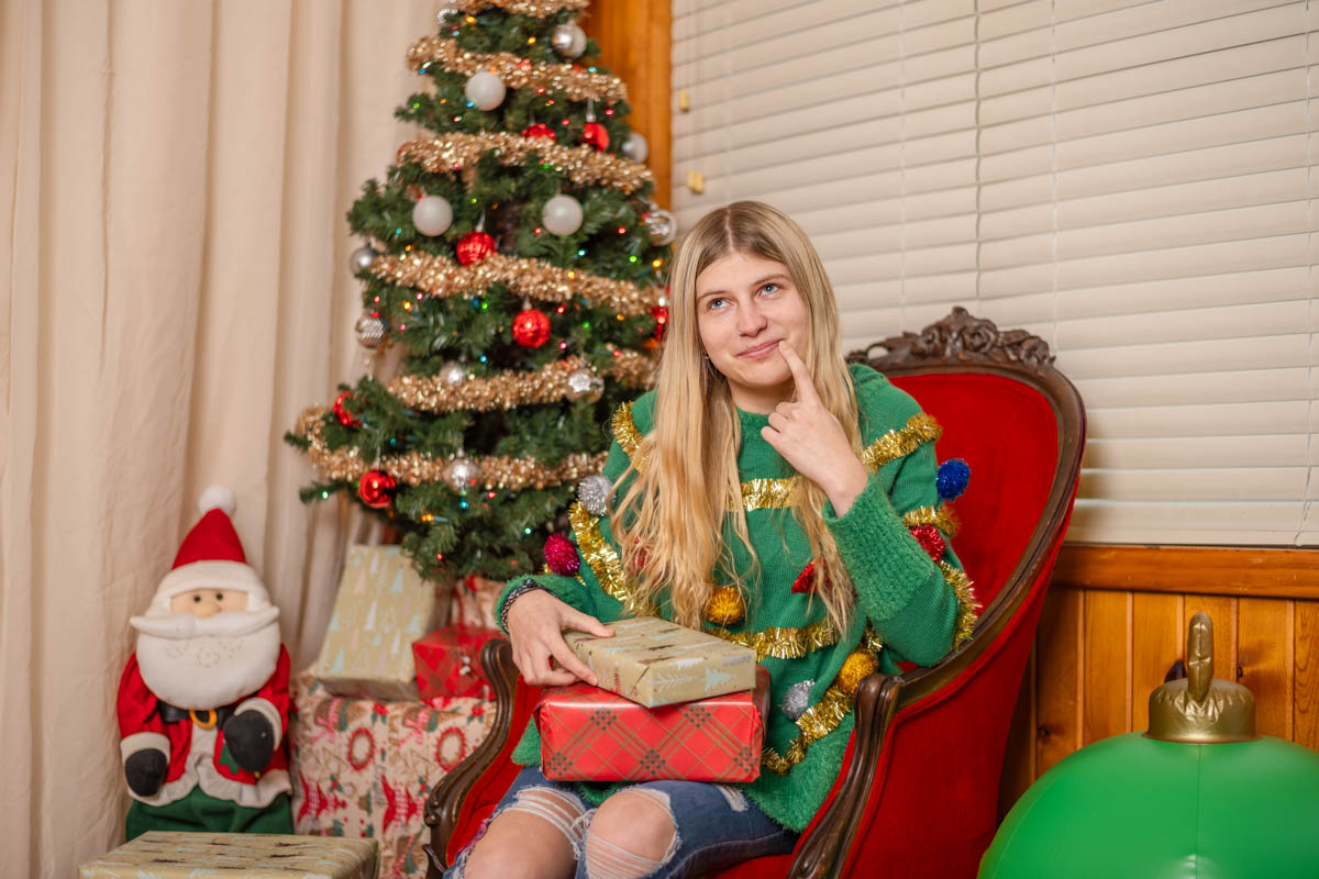 A person sitting in a chair wearing a festive sweater, holding gifts. A decorated Christmas tree and Santa decoration are in the background.