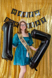 Young woman in a blue dress and tiara holds large black "17" balloons. She wears a "Birthday Queen" sash. The background is gold with a "Happy Birthday" banner.