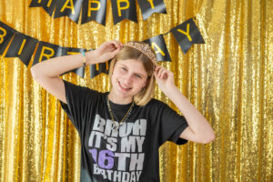 A person smiles while wearing a tiara in front of a gold sequin background with "Happy Birthday" banner. Their shirt reads "Bruh It's My 16th Birthday.