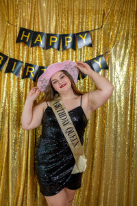 Woman in a black sequin dress and pink cowboy hat stands in front of a gold backdrop with "Happy Birthday" banner, wearing a sash that reads "Birthday Queen.