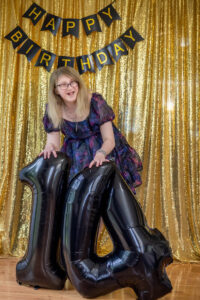 A person smiles while holding black number 14 balloons. A "Happy Birthday" banner hangs on a gold sequin backdrop.