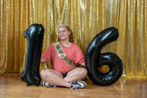 Person wearing a "Birthday Queen" sash and tiara sits on the floor, holding black number balloons "1" and "6" against a gold sequin backdrop.