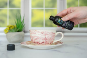 A hand pours CBD oil from a bottle into a teacup on a saucer, with a small plant in the background.