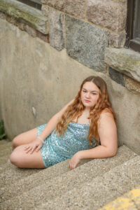 A female student model seated on outdoor steps against a stone wall, wearing a sparkly teal dress.