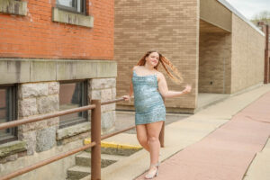 Female high school student in a sparkly blue dress poses confidently on a sidewalk, with brick buildings in the background.