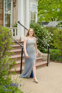 A female high school senior in a silver dress stands on an outdoor staircase in front of a building, surrounded by greenery and flowers.