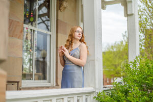 A female high school senior in a metallic dress stands on a porch, gazing to the side, with a window and greenery in the background.