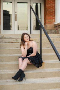 A female high school student in a black lace dress and boots sits on outdoor steps, resting her chin on her hand.