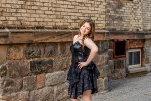 A female high school student in a black sequined dress stands against a brick and stone wall, smiling with one hand on their hip.