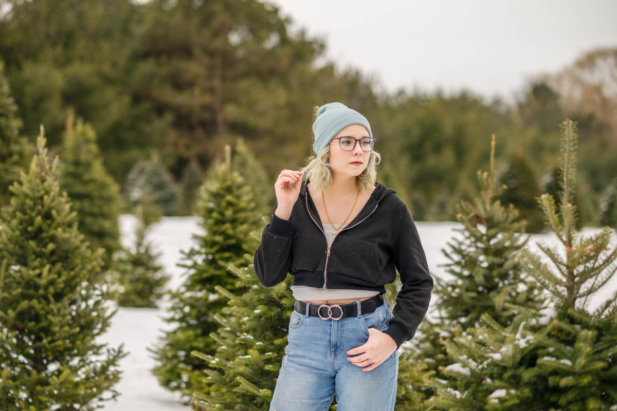 A high school senior in a blue beanie and glasses stands among snow-covered evergreen trees, wearing a black jacket and jeans.