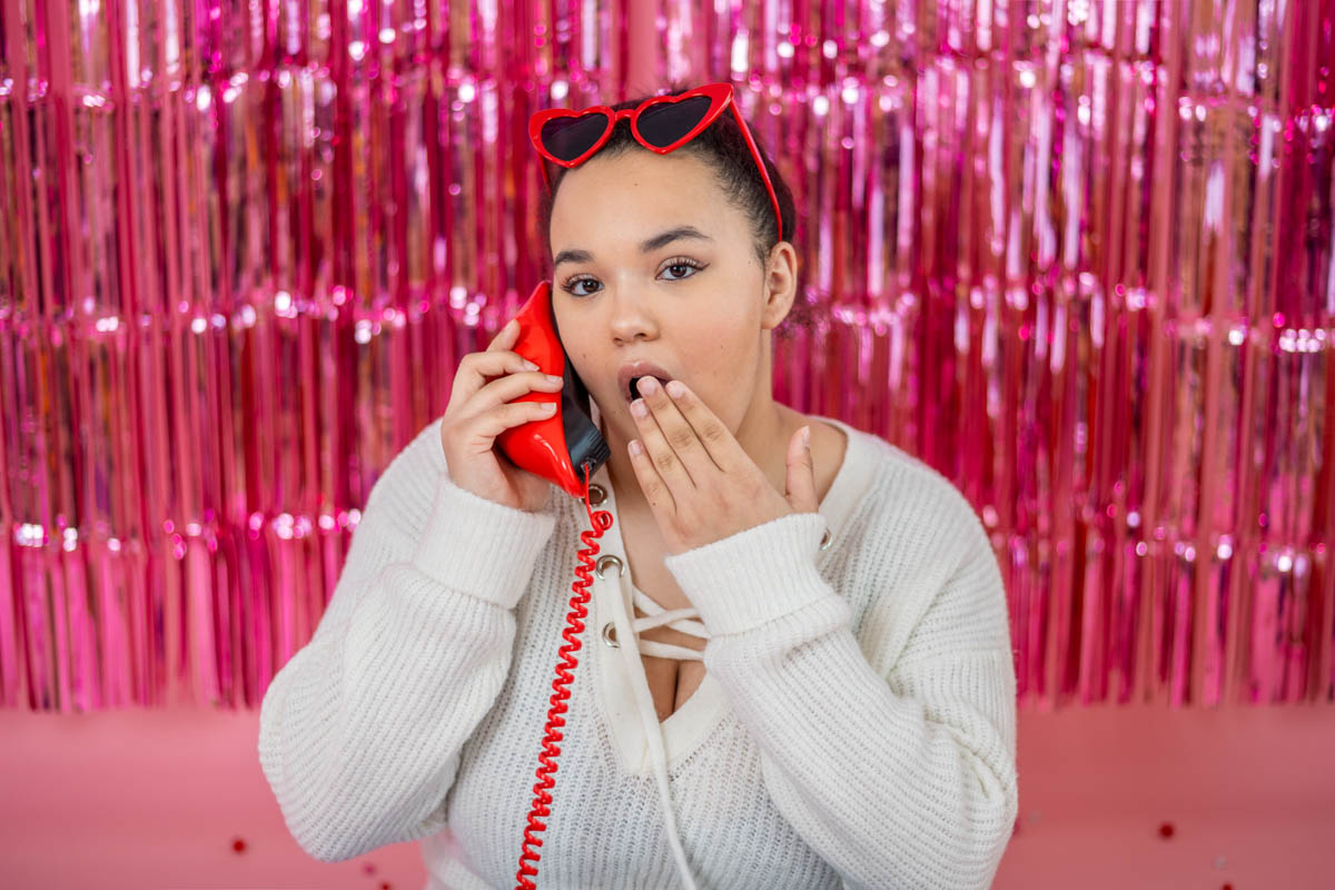 A Female High School Senior with a surprised expression holds a red phone to their ear. They are wearing a white sweater and red sunglasses in front of a pink tinsel backdrop for a Valentine's Day photo shoot.