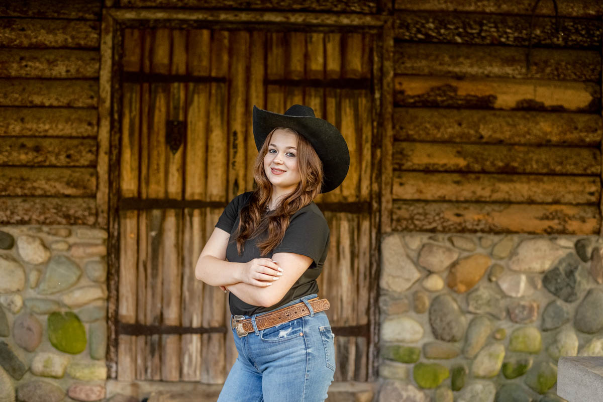 A female high school student wearing a cowboy hat stands with arms crossed in front of a rustic wooden door and stone wall.